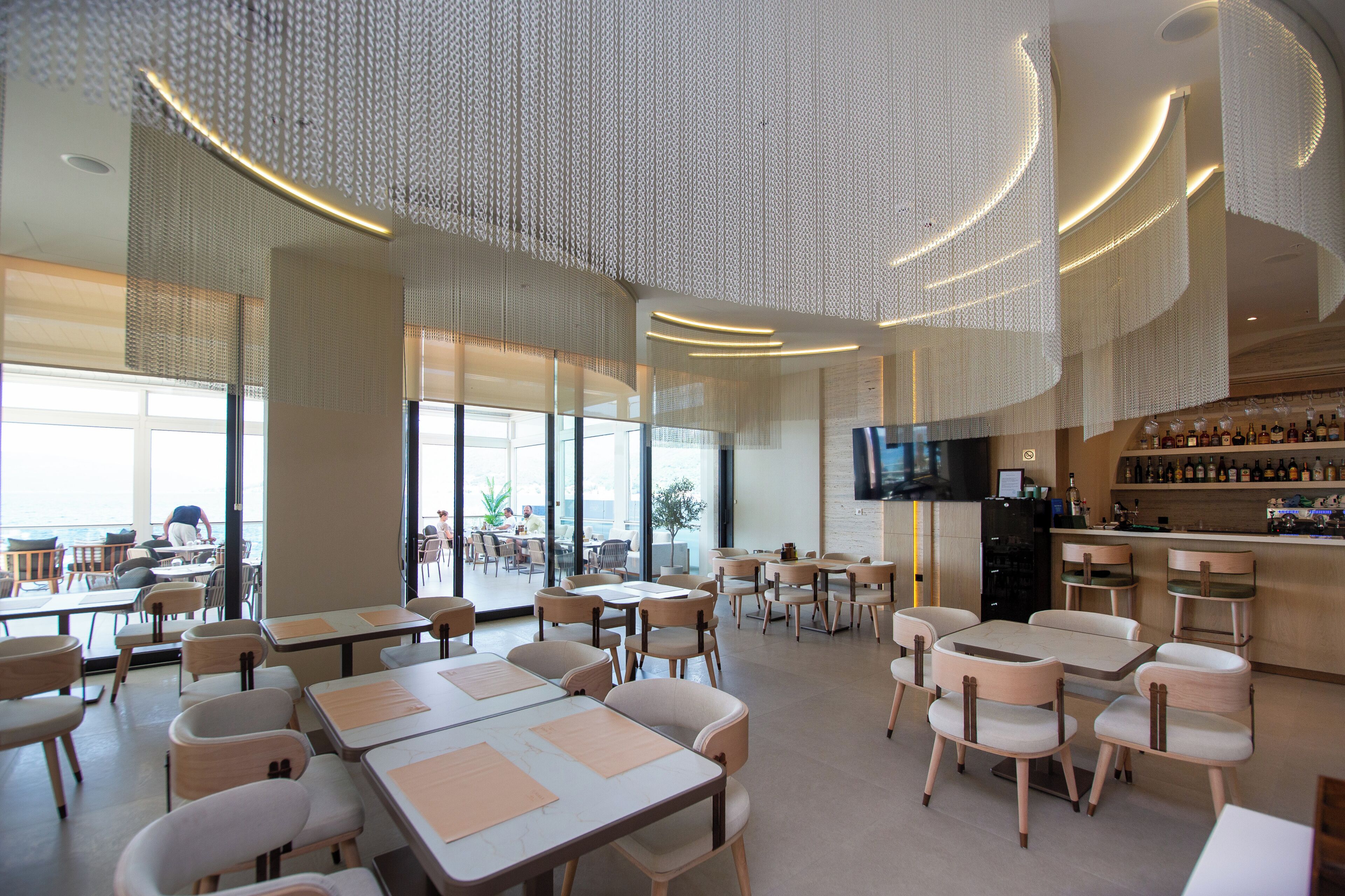 A modern restaurant with a bar area, large windows, and a ceiling with a unique light fixture.