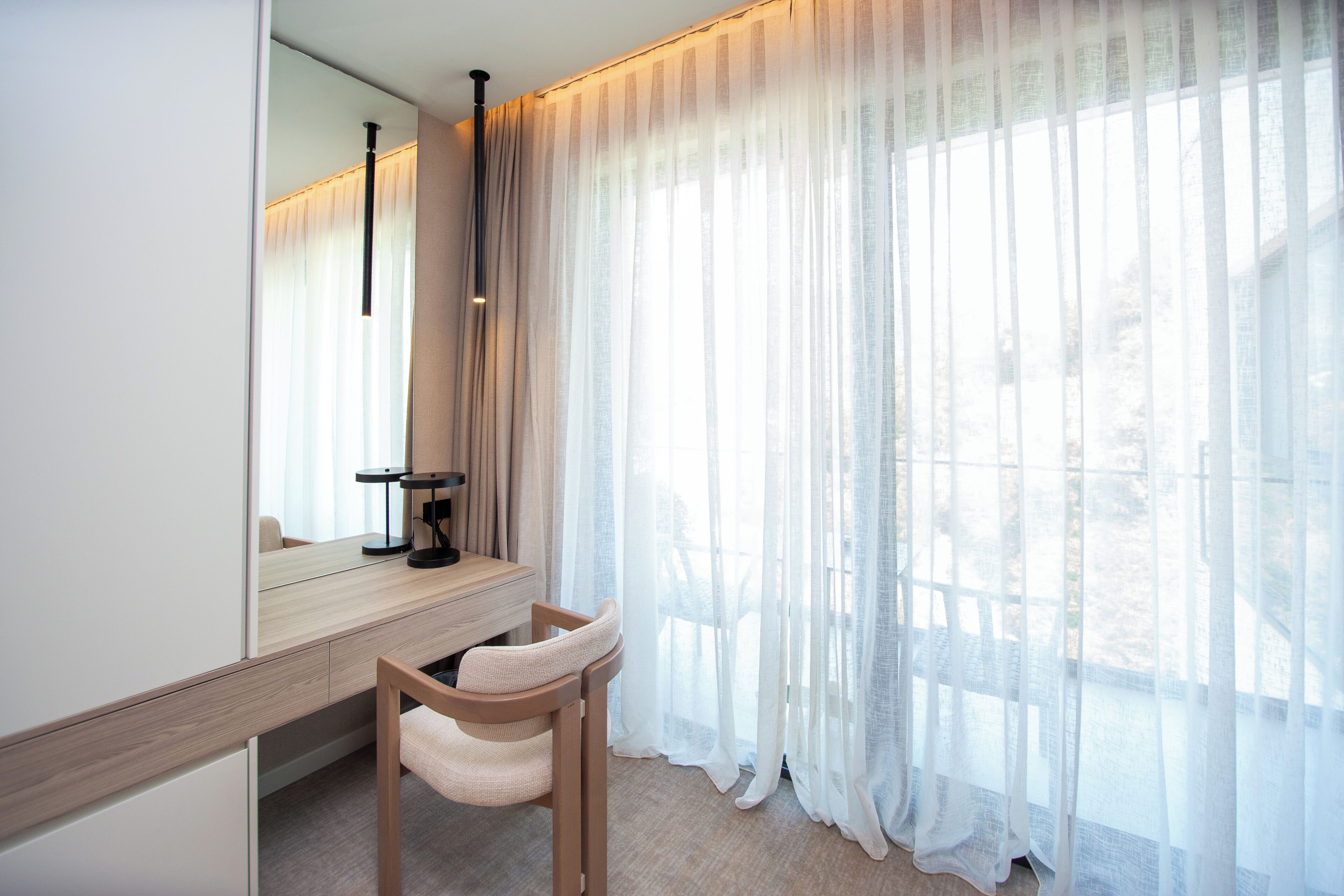 Una habitación de hotel moderna con un espejo grande, un escritorio de madera, una silla y cortinas traslúcidas.