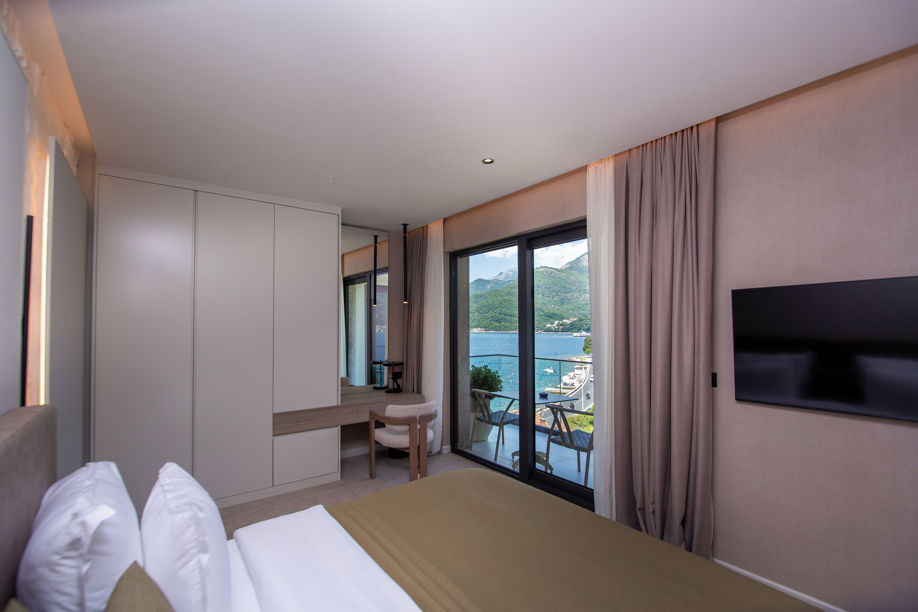 Habitación de hotel con cama, escritorio, silla, televisor y balcón con vista al mar y a las montañas.