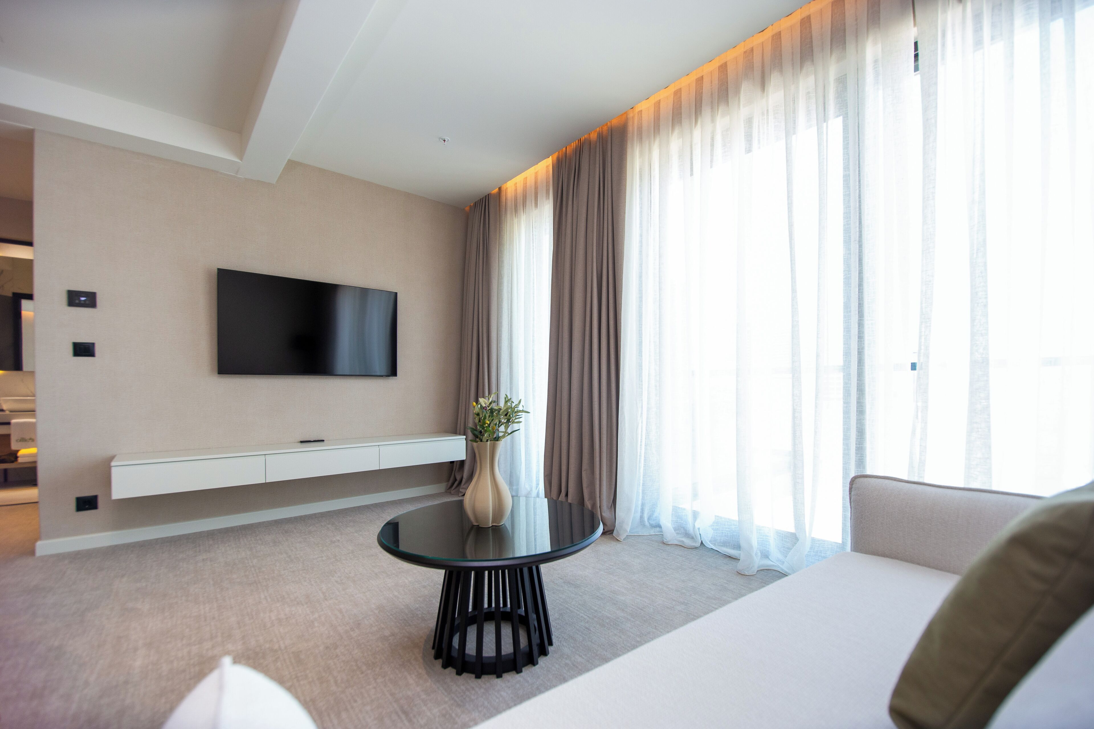 Een moderne hotelkamer met een flatscreen televisie aan de muur, een rond glazen tafeltje met een vaas, een bank en grote ramen met dunne gordijnen.