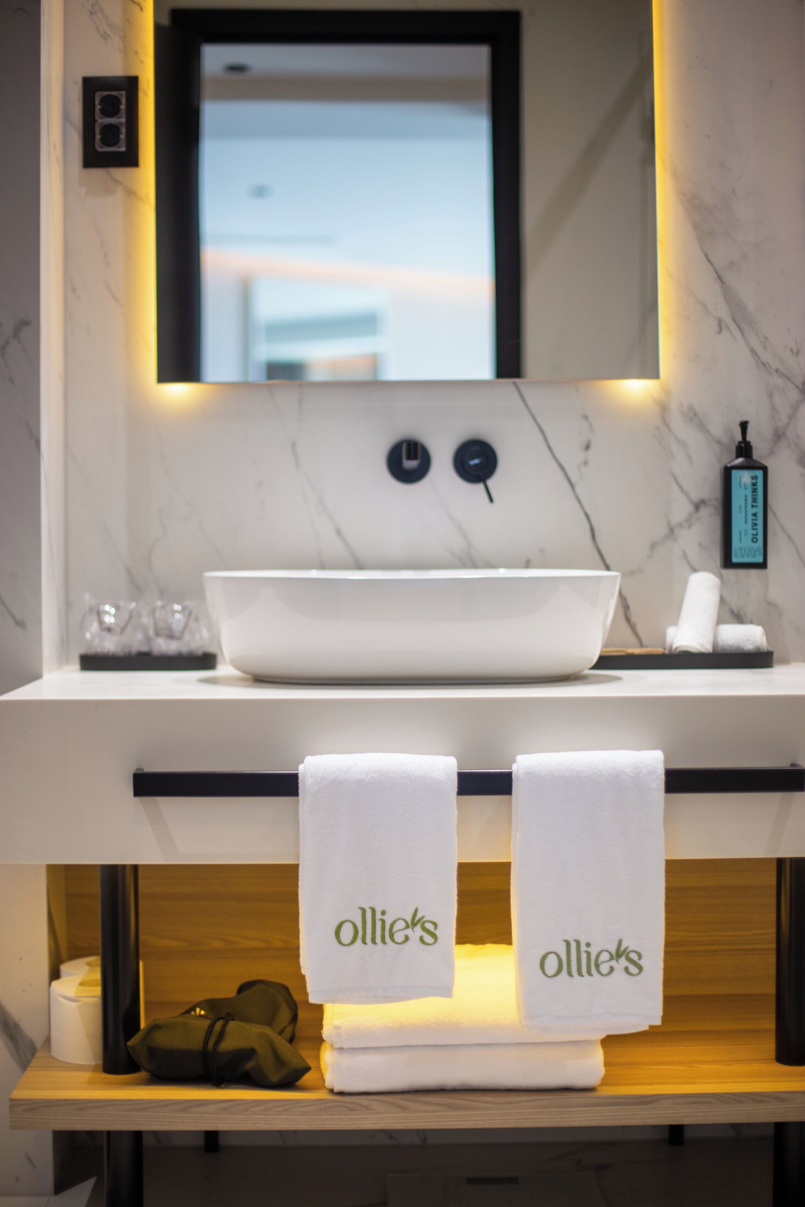 Een badkamer met een witte wasbak, twee handdoeken met 'ollies'-branding, een spiegel en een fles handzeep.