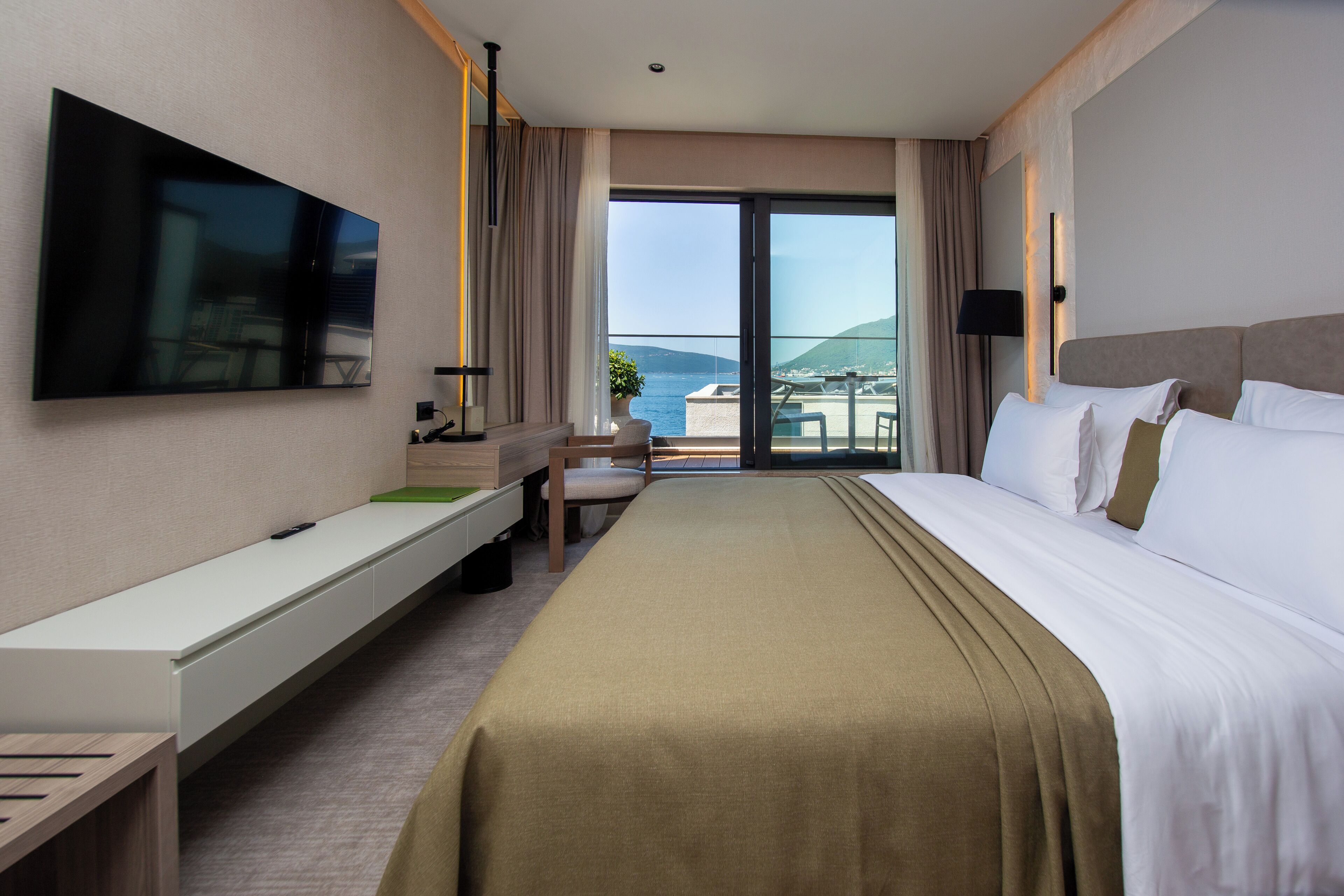 A hotel room with a bed, a TV, a desk, and a view of the sea.