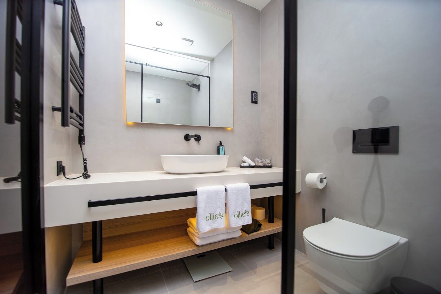 A modern bathroom with a white sink, mirror, and toilet. There are towels stacked on a shelf and a shower area visible.