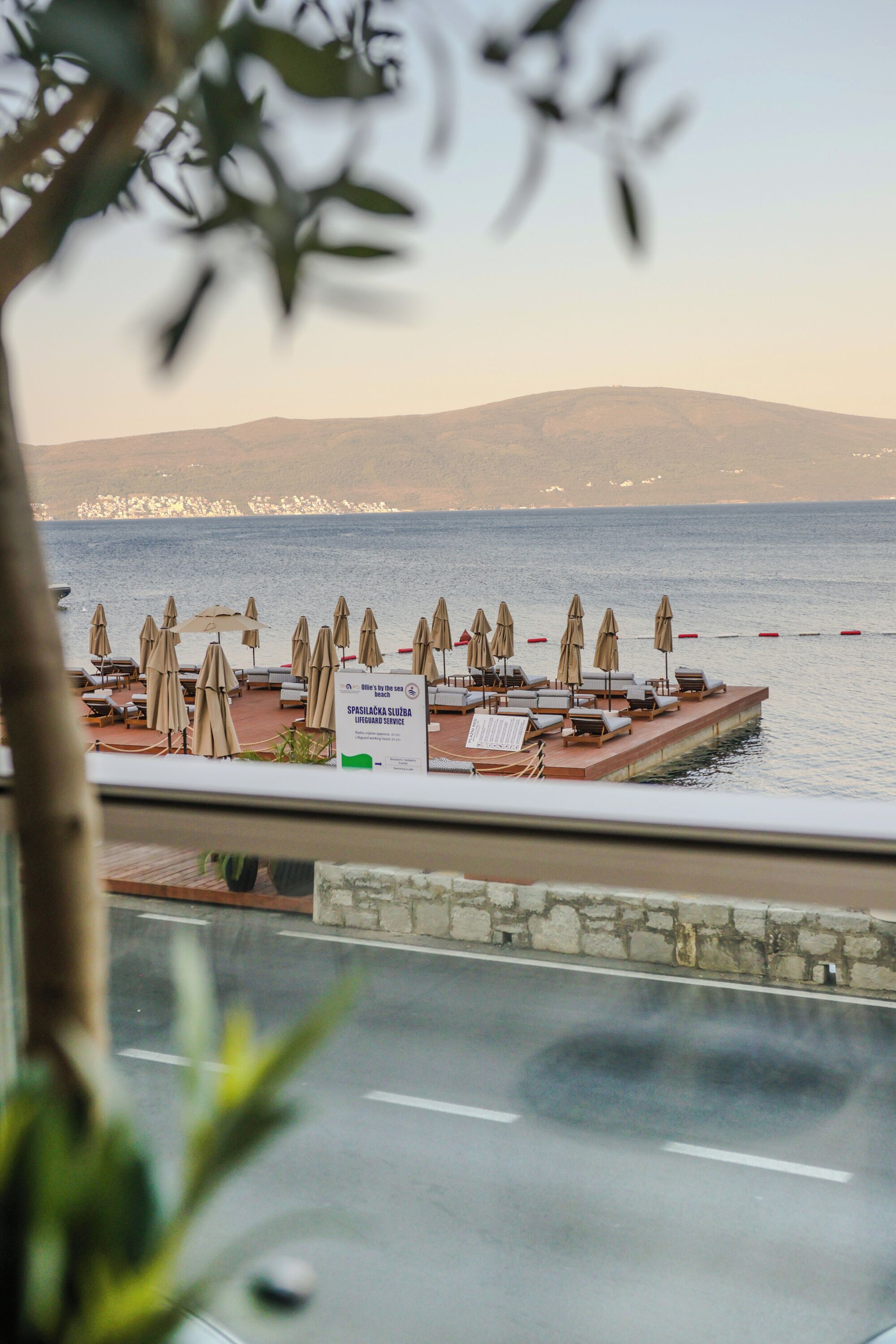A waterfront view with beach umbrellas and lounge chairs, a lifeguard service sign, and a distant cityscape.