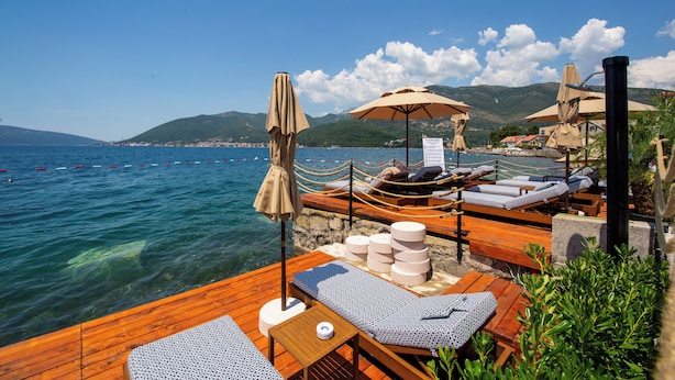 Une terrasse en bois avec des chaises longues et des parasols, donnant sur une étendue d’eau avec des montagnes en arrière-plan.