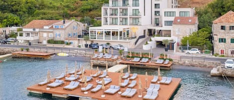 Un hôtel situé au bord de l’eau, avec plusieurs étages, une jetée dotée de chaises longues et un paysage montagneux en toile de fond.