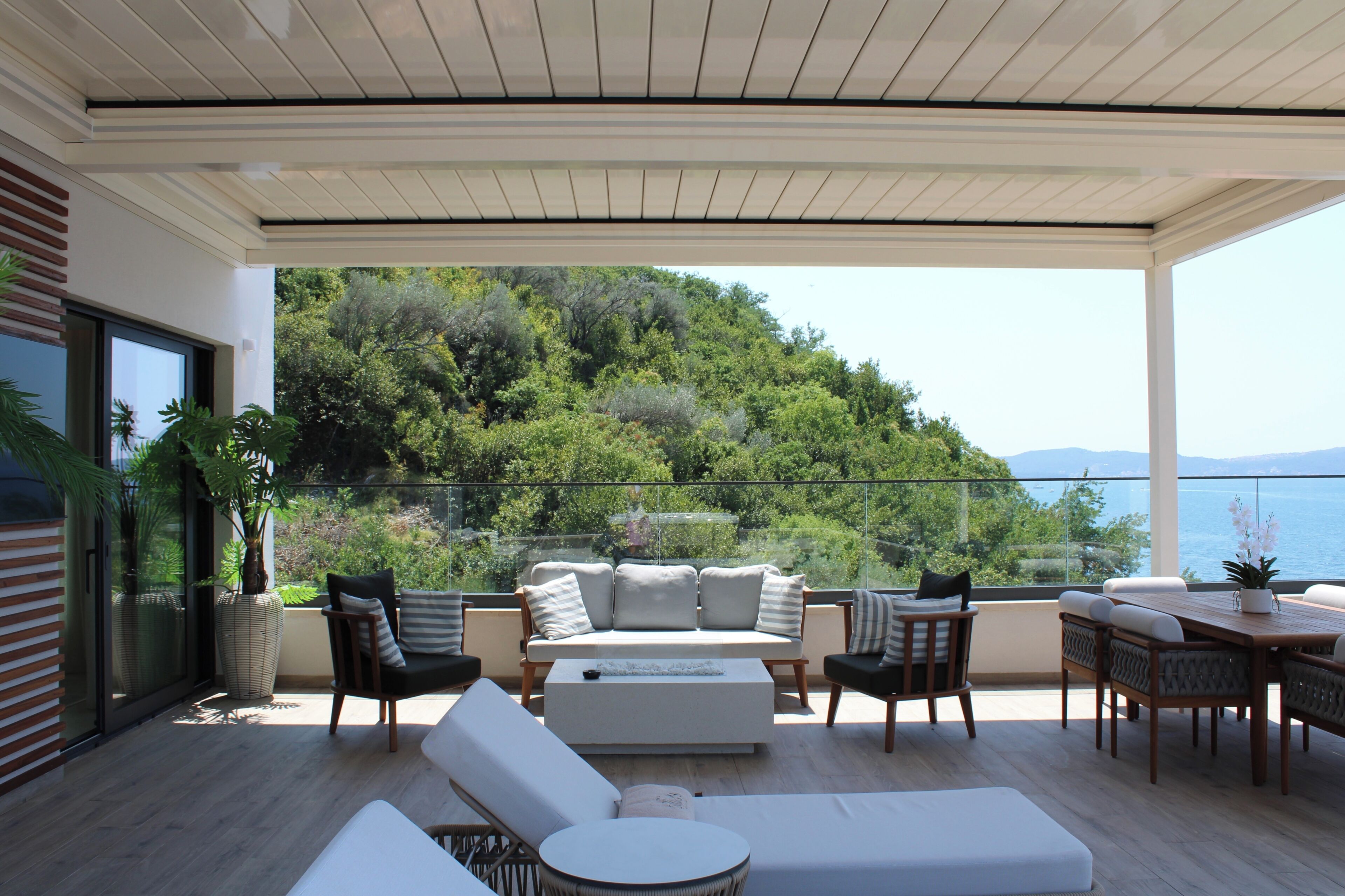 A modern outdoor living space with a sofa, chairs, and a table, overlooking a scenic hillside and body of water.