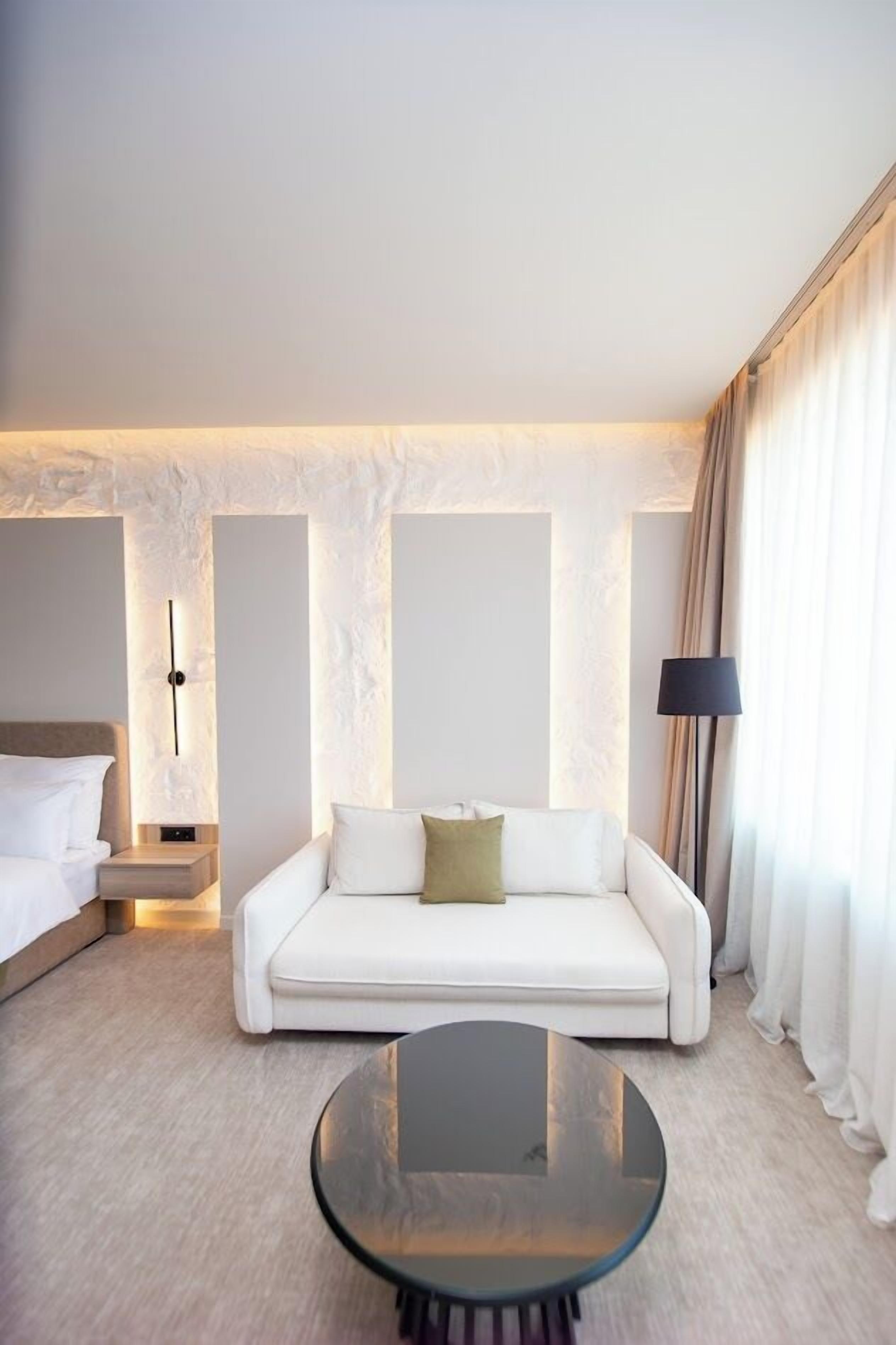 A modern hotel room with a white sofa, a round glass coffee table, a bed with white linens, and a lamp.