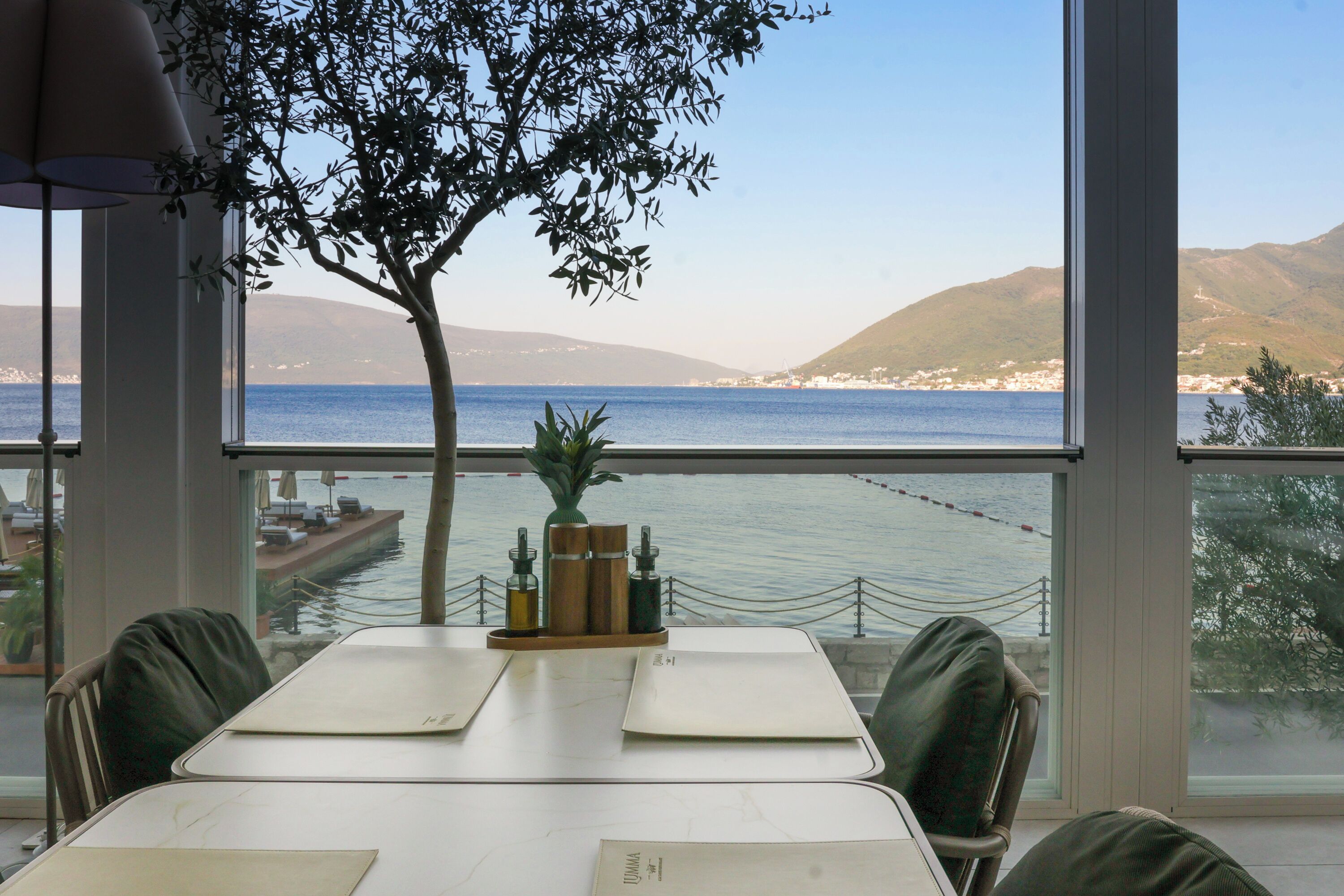 A dining table with chairs, a vase with plants, and a view of the sea and hills through large windows.