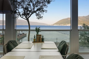 A dining table with chairs, a vase with plants, and a view of the sea and hills through large windows.