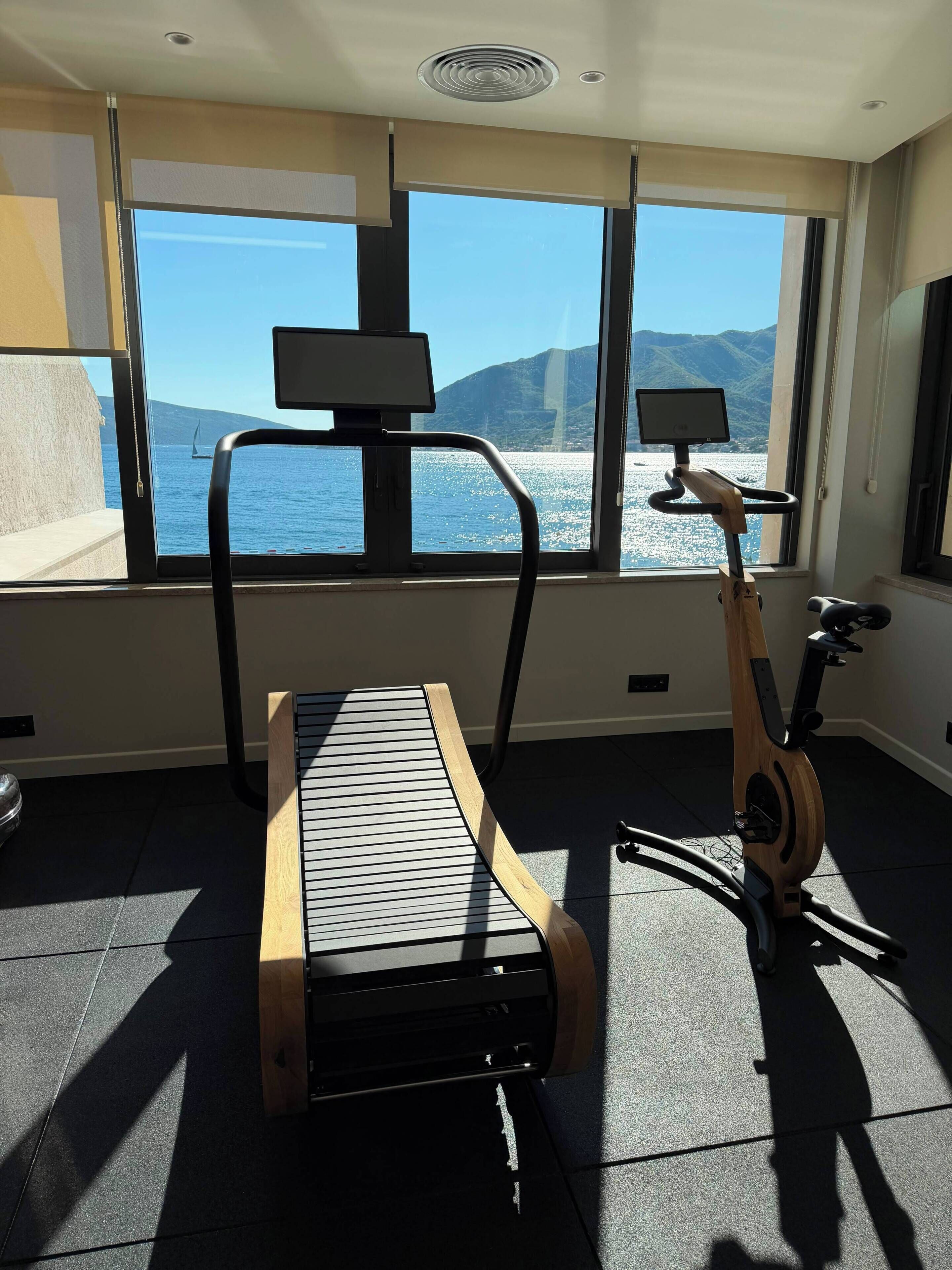 Ein Raum mit einem stationären Fahrrad, einem Laufband und einem großen Fenster mit Blick auf das Meer und die Berge.