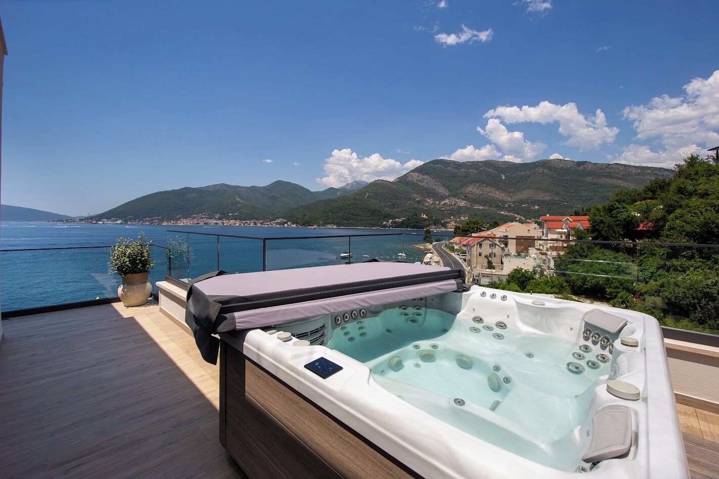 A deck with a hot tub and a potted plant, overlooking a body of water and a mountainous landscape.