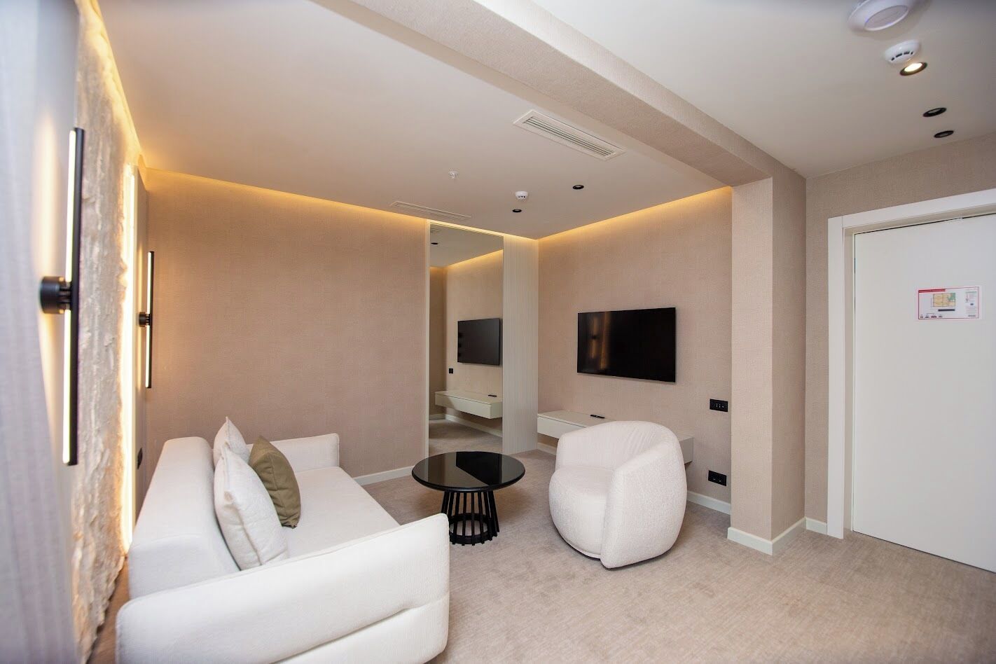 A modern hotel room with a white sofa, a round coffee table, a white armchair, a flat-screen TV mounted on the wall, and a door with a security card reader.