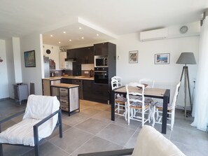 Une cuisine moderne avec des meubles de couleur sombre, un coin repas avec une table et des chaises, et un salon avec un canapé et une lampe.