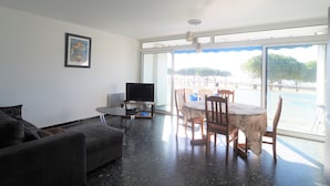 A living room with a sofa, a TV, a dining table with chairs, and a large window with a view of trees and a pool.