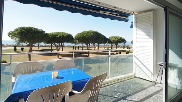 A balcony with a table and chairs, overlooking a beach with trees and the sea.