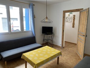 A room with a sofa, a table with a leaf-patterned tablecloth, a television on a stand, a hanging light fixture, and a door with a wooden frame. - Fisherman's house with AC, city - 6 guests (Le Grau-du-Roi)
