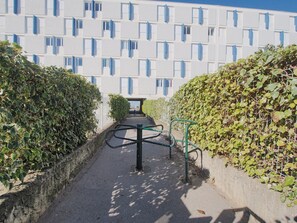 A pathway leading to a building with a modern architectural design, featuring a geometric pattern of windows and a hedge-lined entrance. - Apartment with AC and parking - 6 guests, steps from the beach (Le Grau-du-Roi)