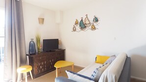 Una sala de estar con un sofá, un televisor sobre un mueble de madera, una mesita y una decoración de veleros en la pared.