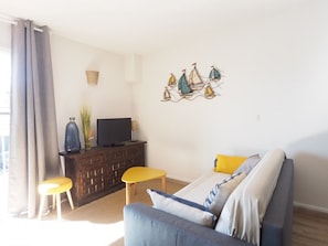 A living room with a sofa, a television on a wooden cabinet, a small table, and a wall decoration of sailboats. - Apartment with AC and parking - 6 guests, steps from the beach (Le Grau-du-Roi)