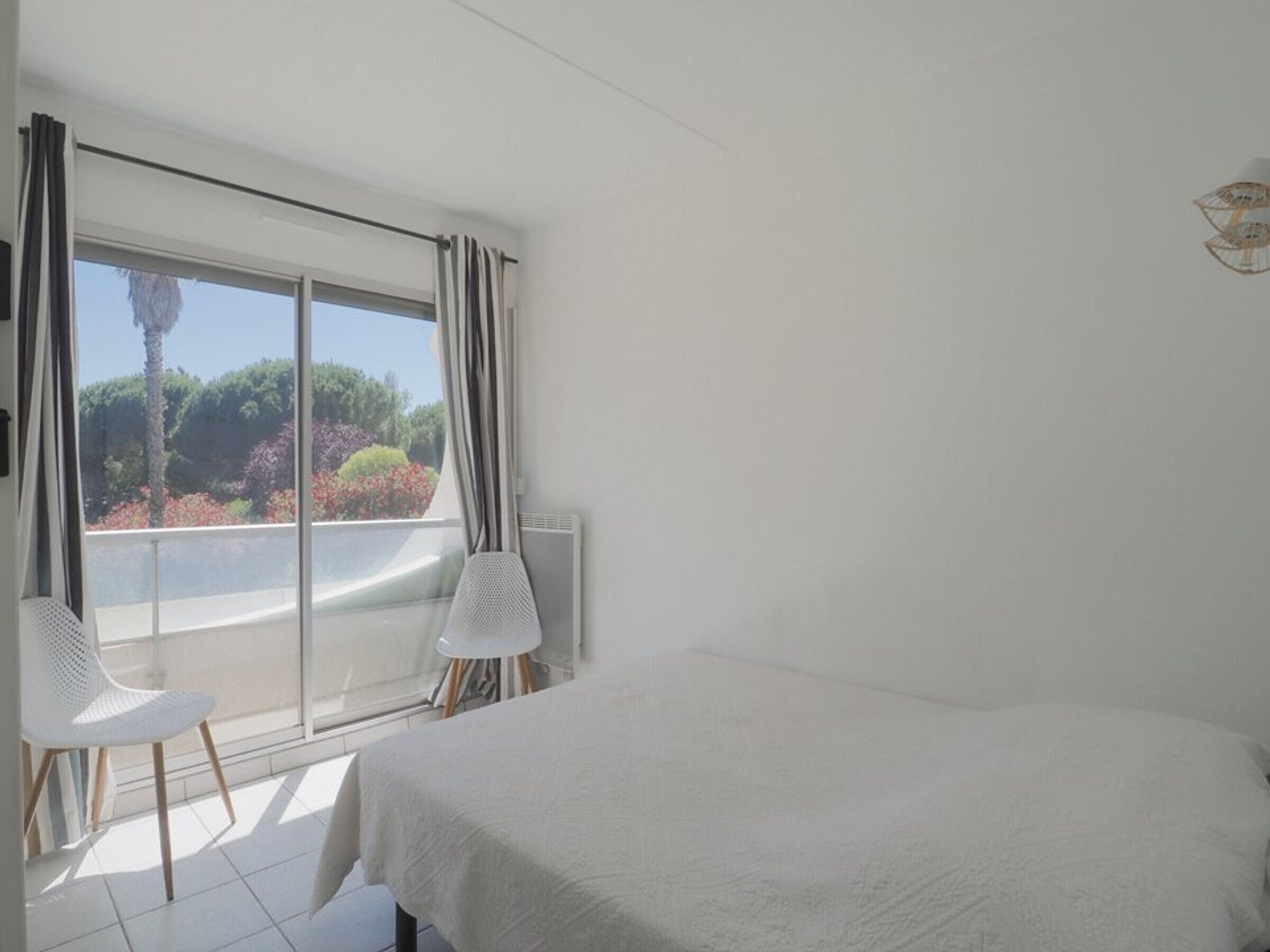 Un dormitorio con cama, dos sillas, un ventanal con cortinas y vistas a zonas verdes.