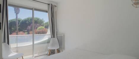 Un dormitorio con cama, dos sillas, un ventanal con cortinas y vistas a zonas verdes.