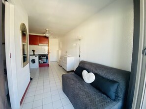A living room with a sofa, a heart-shaped pillow, a ceiling fan, and a kitchen in the background. - Studio cabin near beach - 5 guests (La Grande-Motte)