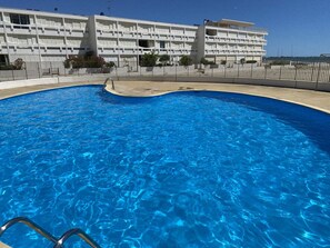 A swimming pool with a metal ladder, surrounded by a fence, and a white building with multiple balconies in the background. - Studio at Boucanet, parking - 4 guests (Le Grau-du-Roi)