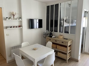 A modern, minimalistic room with a white table, chairs, a flat-screen TV, a fan, and a large window with a view. - Spacious apartment with pool - 6 guests (La Grande-Motte)