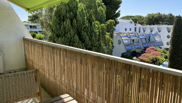 Un balcon doté d’une balustrade en osier, d’une chaise en rotin et offrant une vue sur un immeuble aux balcons bleus et sur un arbre.