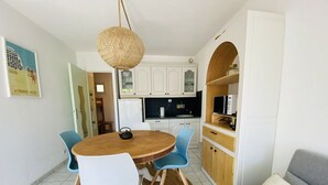 Un coin repas avec une table ronde en bois, deux chaises, un luminaire suspendu en fibres tressées et, au fond, une cuisine.