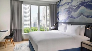 Habitación de hotel con una cama grande, un escritorio y vistas a edificios de la ciudad a través de amplias ventanas.