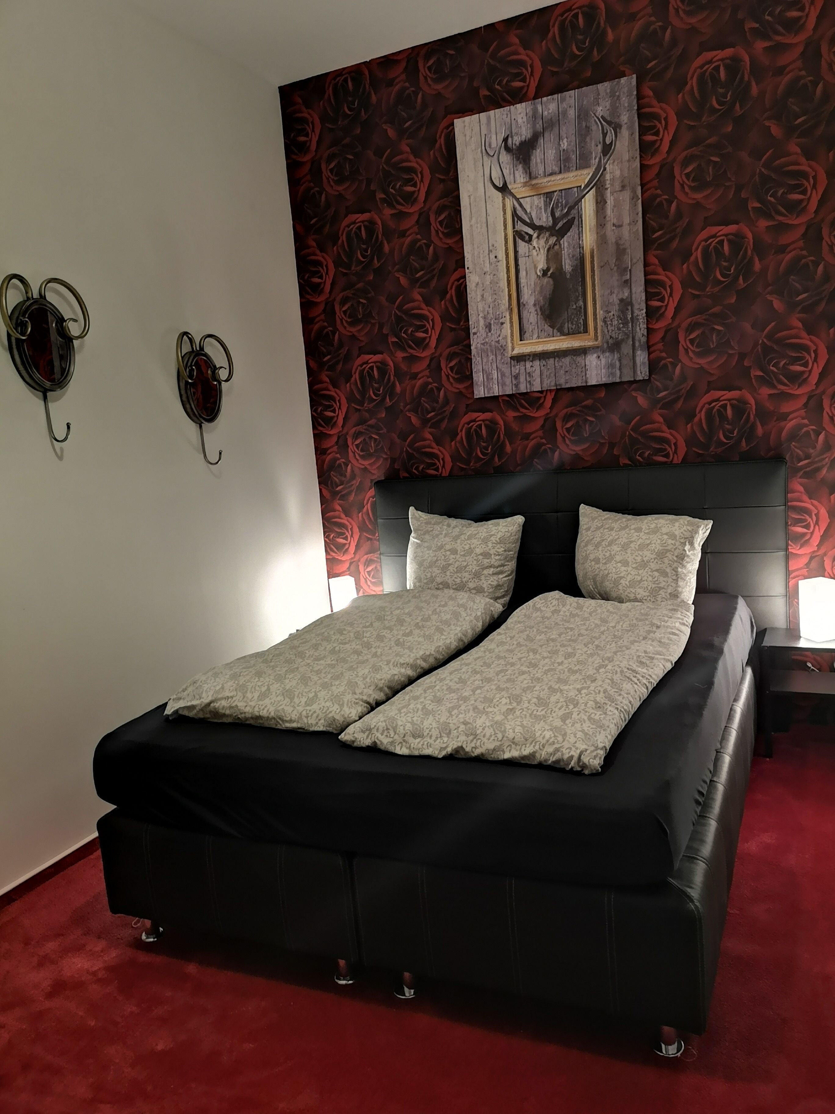 Une chambre avec un lit noir, deux appliques murales, une moquette rouge et un papier peint à motifs floraux.