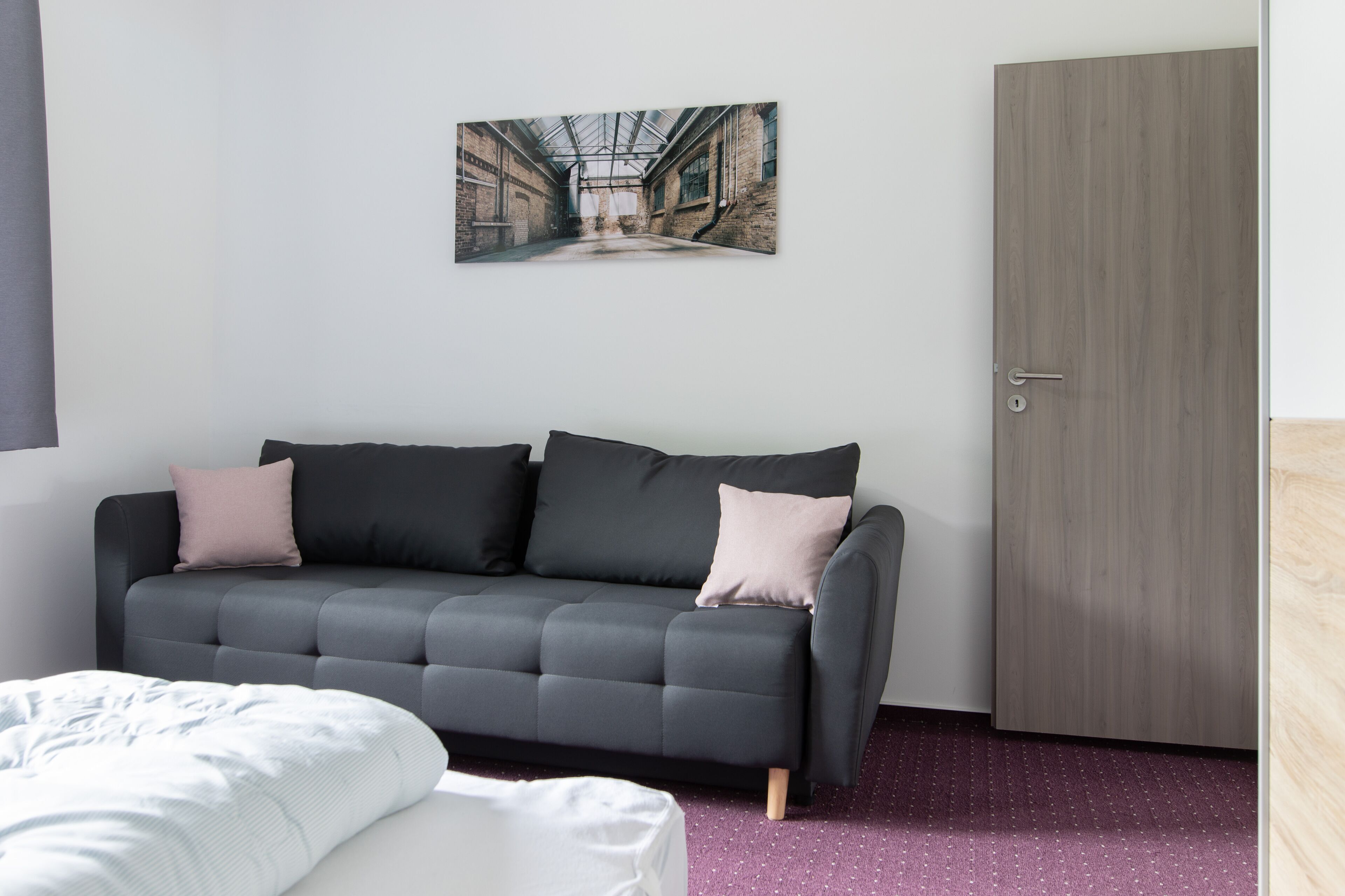 Une chambre moderne avec un canapé gris, un lit, une porte en bois et un tableau représentant un couloir industriel, accroché au mur.