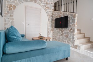 A living room with a blue sofa, a stone wall, a flat-screen TV, and a staircase. - Aegean Nest | 2-Bedroom House with Castle Views (Mitilini)
