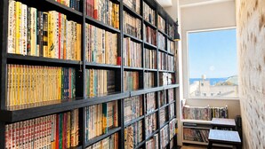 A library with bookshelves filled with books, a window with a view, and a small table with chairs. - ArtBar & GuestHouse ennova - Hostel (Atami)