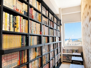 A library with bookshelves filled with books, a window with a view, and a small table with chairs. - ArtBar & GuestHouse ennova - Hostel (Atami)