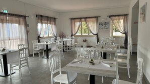 A dining area with white chairs and tables, large windows with sheer curtains, and a clean, modern interior.