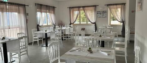 A dining area with white chairs and tables, large windows with sheer curtains, and a clean, modern interior.