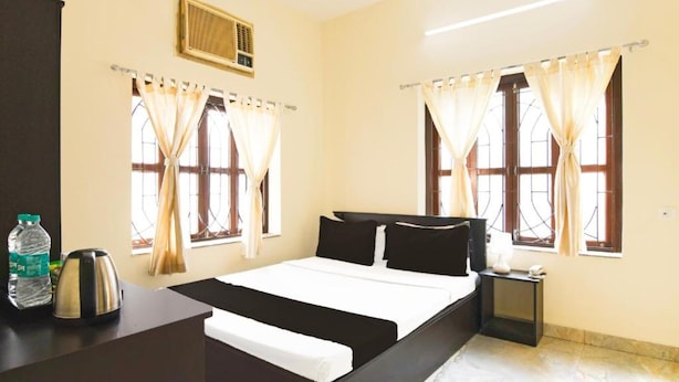 A hotel room with a bed, a nightstand, a kettle, a bottle of water, and curtains on the windows. - Kiya Kedia Niketan Salt Lake (Barasat)
