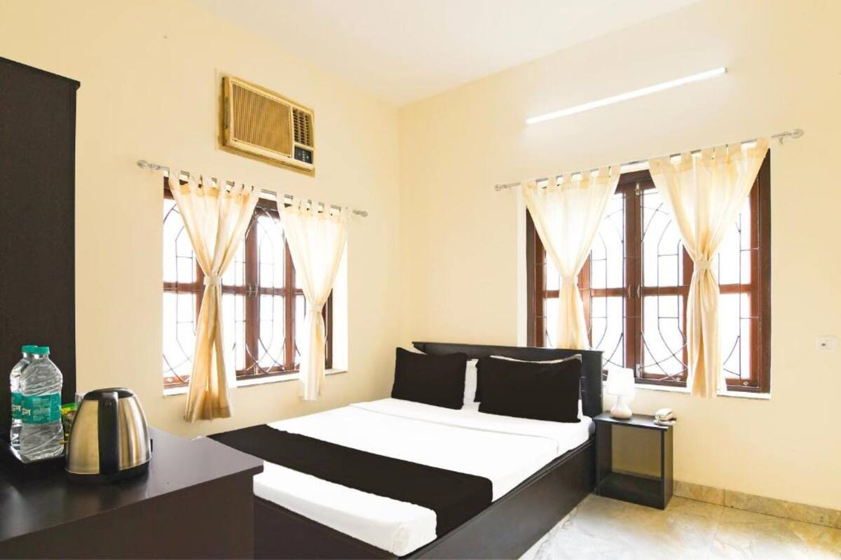 A hotel room with a bed, a nightstand, a kettle, a bottle of water, and curtains on the windows.