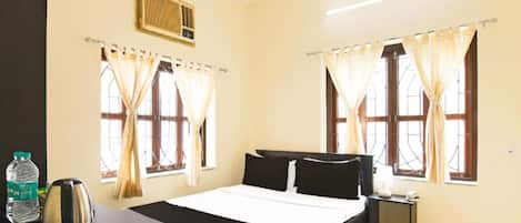 Habitación de hotel con una cama, una mesita de noche, un hervidor de agua, una botella de agua y cortinas en las ventanas.