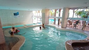 An indoor swimming pool with a tiled interior, a visible sign on the wall, and a seating area outside the pool. - Apartment for 4 people in Bombas + swimming pools + gym + sauna + games rooms (Bombinhas)