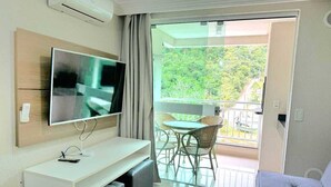 A modern room with a flat-screen TV, a white console table, a sofa, a balcony with a table and chairs, and a view of greenery. - Apartment for 4 people in Bombas + swimming pools + gym + sauna + games rooms (Bombinhas)