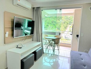 A modern room with a flat-screen TV, a white console table, a sofa, a balcony with a table and chairs, and a view of greenery. - Apartment for 4 people in Bombas + swimming pools + gym + sauna + games rooms (Bombinhas)