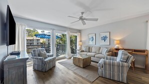 A spacious living room with a large TV, a ceiling fan, and a view of a balcony with outdoor furniture. - Spacious and modern 3 bedroom villa with stunning lagoon views (Hilton Head Island)