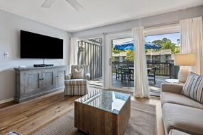 Un salon équipé d’un téléviseur à écran plat, d’une table basse en bois, d’un fauteuil à rayures, d’un canapé et d’une porte-fenêtre coulissante donnant sur une terrasse extérieure avec une table et des chaises.