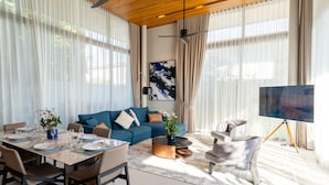 A modern living room with a blue sofa, dining table set, and a large painting on the wall. - Villa Kaya (Thep Krasattri)