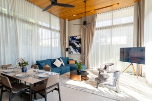 A modern living room with a blue sofa, dining table set, and a large painting on the wall. - Villa Kaya (Thep Krasattri)