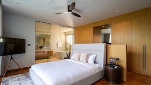A modern bedroom with a large bed, a ceiling fan, a TV, and a bathroom visible through a glass door. - Villa Kaya (Thep Krasattri)